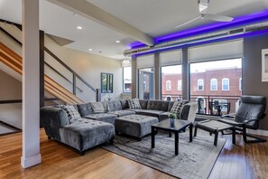 Ein modernes Wohnzimmer mit Ecksofa, Hocker, Couchtisch und Sessel. Eine Treppe mit Holzboden und Geländer ist zu sehen. Große Fenster bieten Blick auf ein Backsteingebäude.