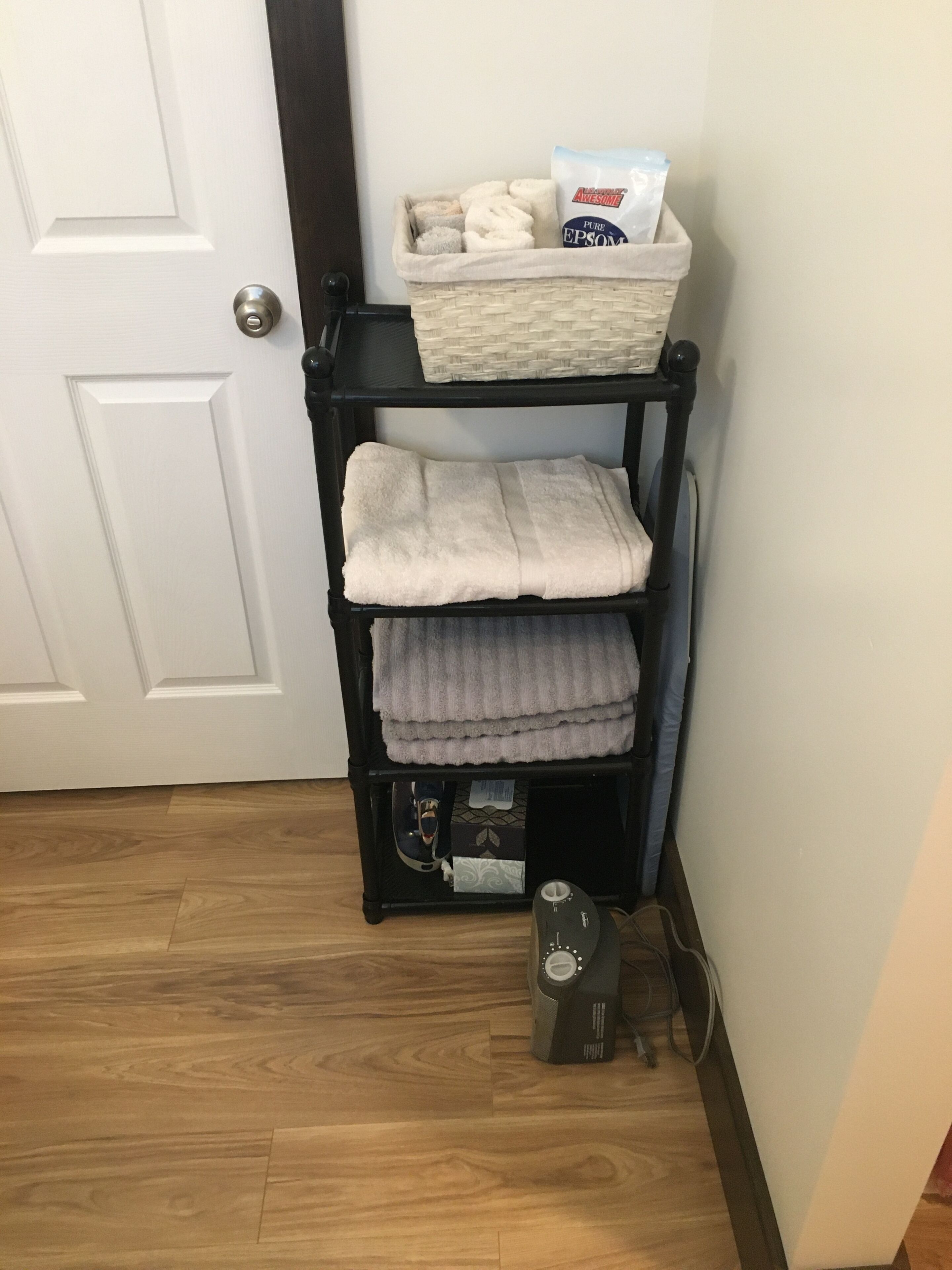 A corner with a black shelf holding folded towels and a basket with a bag of laundry.