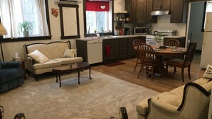 Una sala de estar con un sofá, una mesa de centro y una alfombra estampada. Hay un comedor con una mesa y sillas. La cocina tiene gabinetes de madera y un microondas.