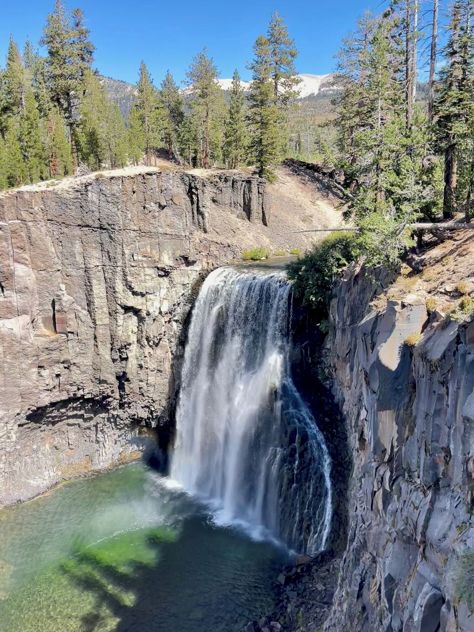 A waterfall cascading over a rocky cliff into a clear pool, surrounded by dense forest and mountains in the background.