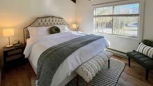 Un dormitorio con una cama grande, una mesita de noche con lámpara, un ventanal con vistas a árboles y un sofá verde.