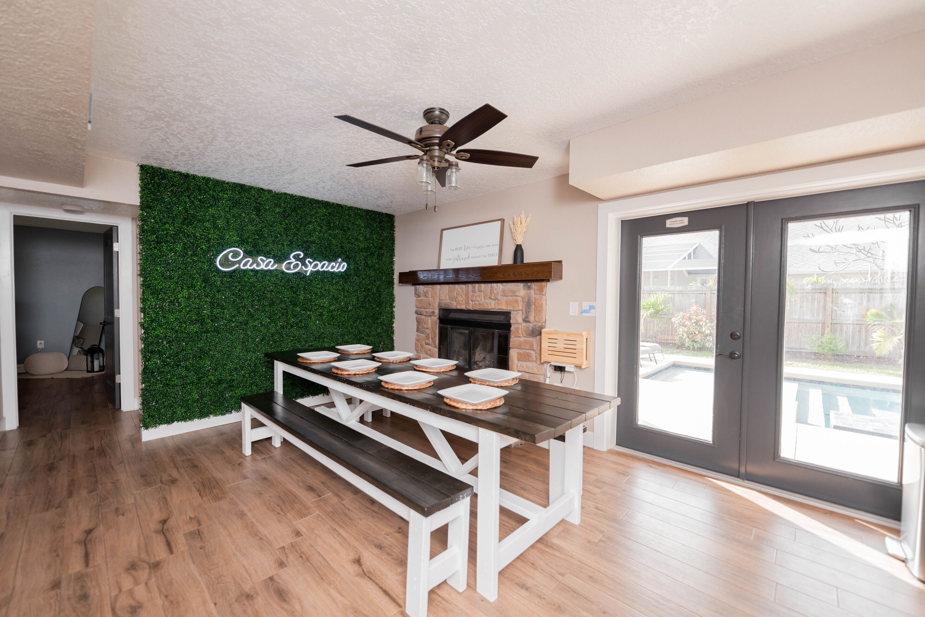A dining area with a wooden table and benches, a green wall with 'Casa Espacio' signage, a fireplace, and a view of a backyard with a pool.