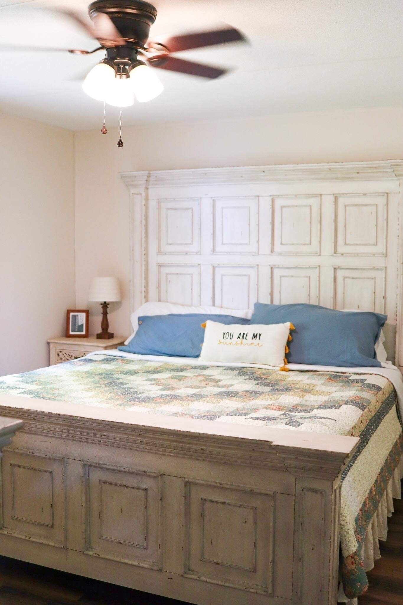 Une chambre à coucher avec une tête de lit blanche, un lit recouvert d’une courtepointe, un ventilateur de plafond et une table de chevet avec une lampe.