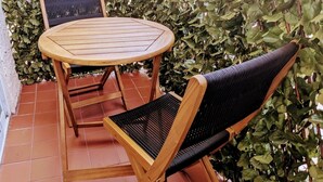 Un tavolo rotondo in legno e due sedie con sedili intrecciati, su un patio piastrellato con uno sfondo di verdeggianti piante.