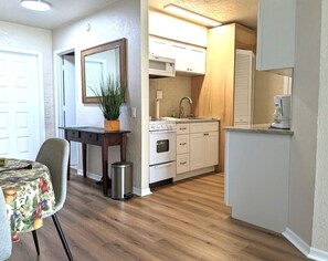 Cocina compacta con armarios blancos, un microondas y un lavamanos. Hay un comedor con una mesa y sillas, y una planta en una mesita.