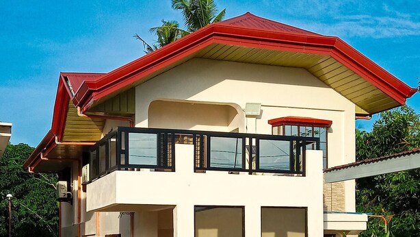 A two-story house with a red roof, multiple windows, and potted plants in the front yard. - La-Rivière: 3-bedroom family-sized house (Lal-lo)