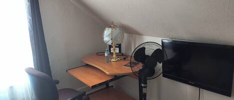 A room with a desk, chair, standing fan, and a mounted TV.