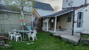 Un patio trasero con mesa y sillas, una zona cubierta pequeña y una casa con antenas parabólicas.