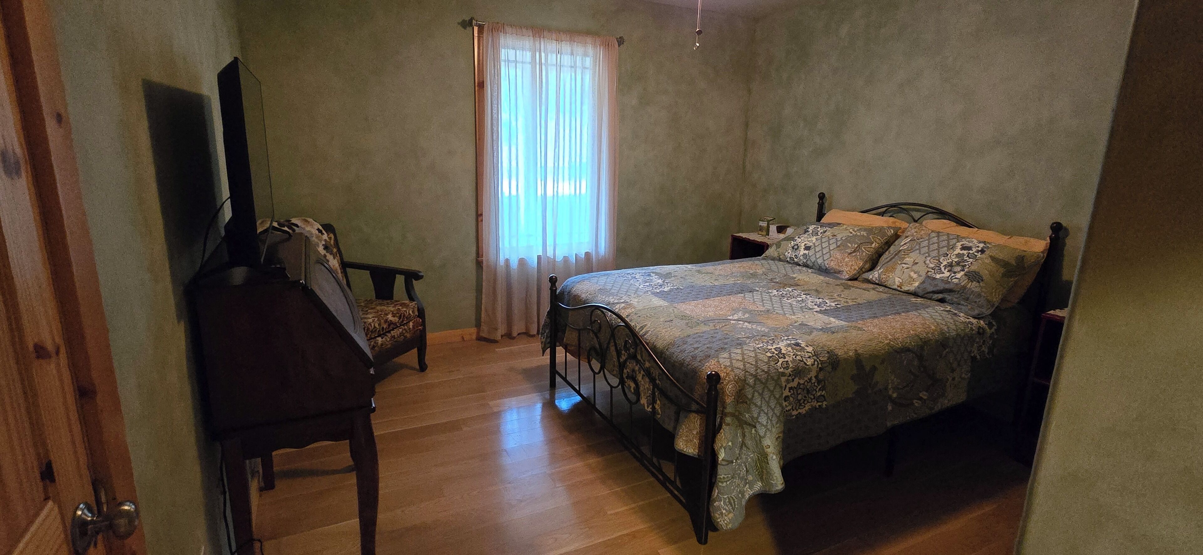 A bedroom with a wooden floor, a metal bed frame, a patterned bedspread, a chair, a small bedside table, and a television on a stand.