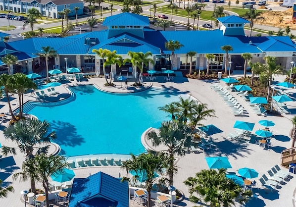 A hotel with a large central pool surrounded by sun loungers and umbrellas, palm trees, and a blue-roofed building.