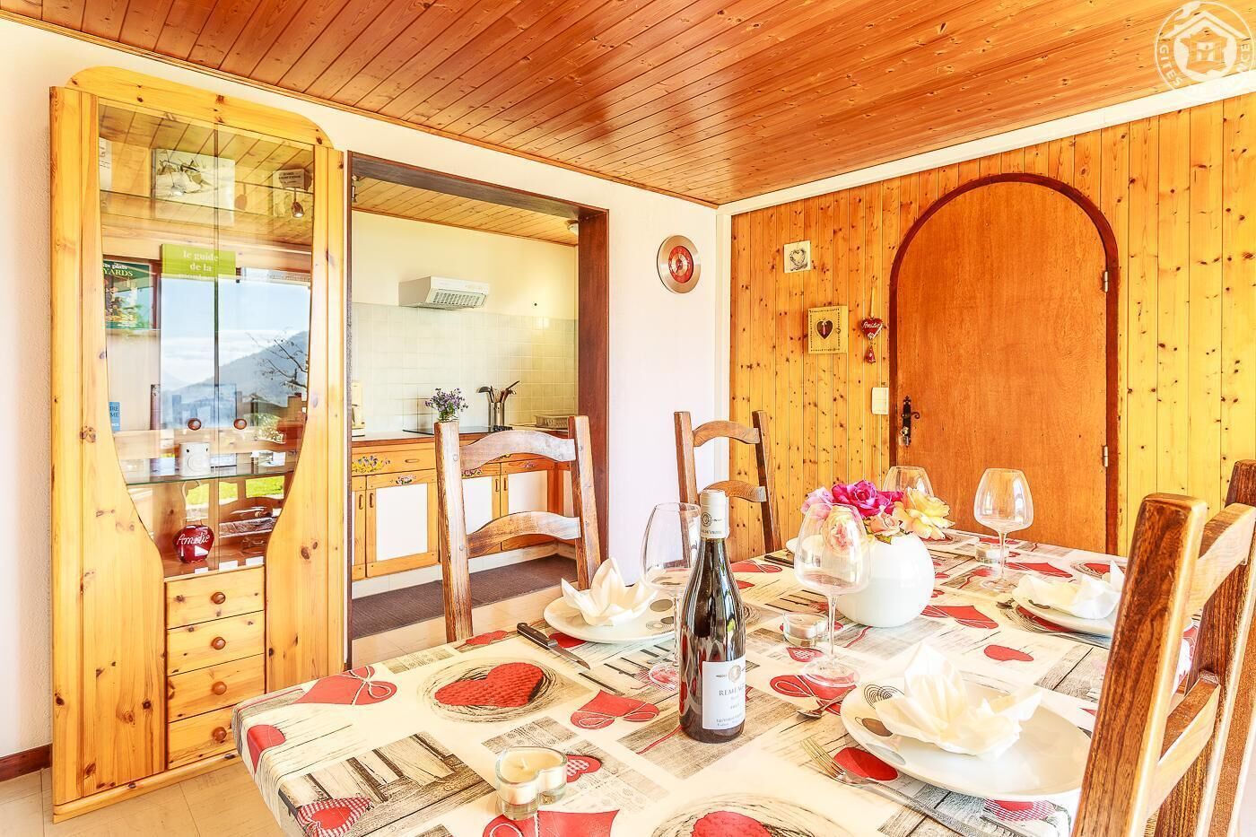 A dining area with a wooden table set for two, a bottle of wine, and a vase with flowers.