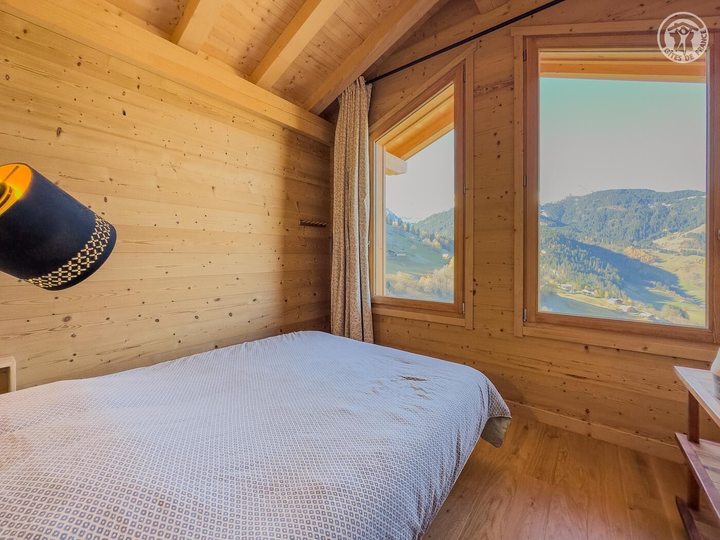 Een houten kamer met een bed, een raam met uitzicht op een vallei en een hanglamp.