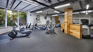 Ein Fitnessstudio mit verschiedenen Trainingsgeräten, einem Holzbüro und großen Fenstern mit Blick auf Grünflächen.