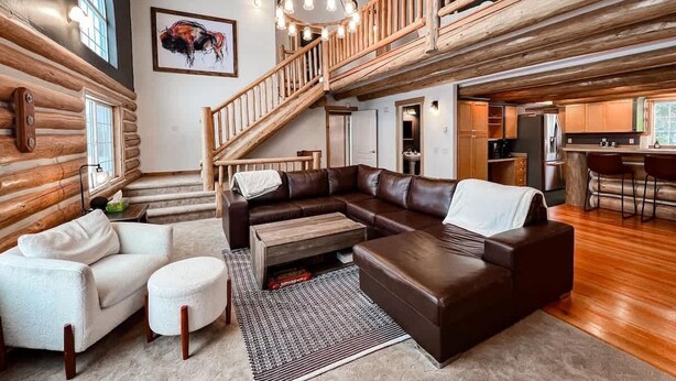A log cabin interior with a large L-shaped sectional sofa, a dining area, and a staircase leading to an upper level.