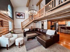 A log cabin interior with a large L-shaped sectional sofa, a dining area, and a staircase leading to an upper level.