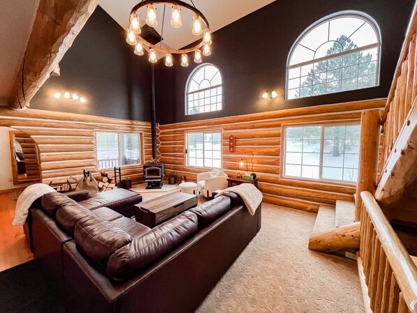 A log cabin interior with a leather sofa, wooden walls, a fireplace, and large windows.