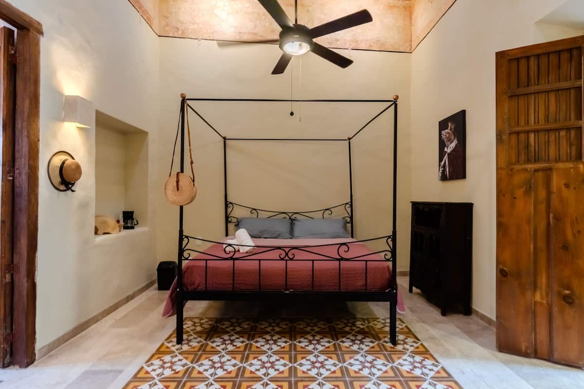 A four-poster bed with a red bedspread, a ceiling fan, a wall-mounted lamp, and a wooden door.