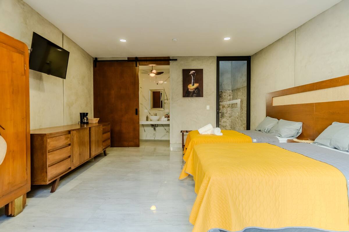 A hotel room with a wooden dresser, a flat-screen TV, two beds with yellow and blue bedding, a wooden headboard, and a marble floor.
