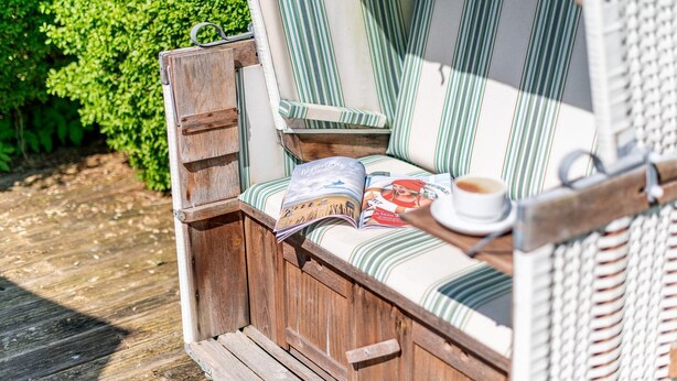 Un banc en bois avec un coussin à rayures, un magazine, une tasse de café et une chaise en osier.