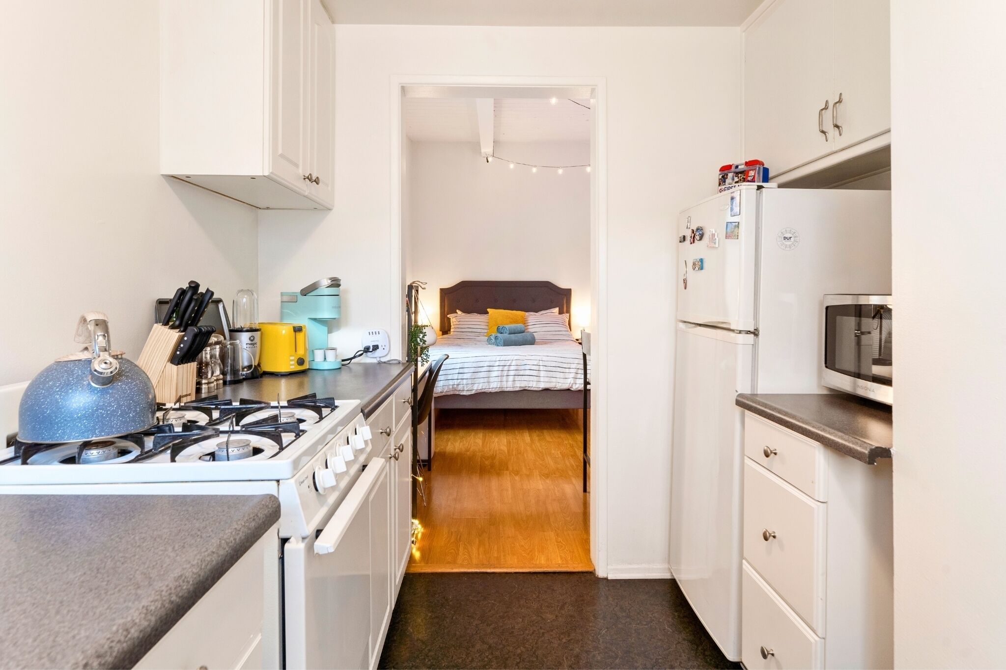 A compact kitchen with a gas stove, a microwave, and a refrigerator. There is a bed with a wooden headboard and a bedside table with a lamp.