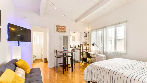 A cozy bedroom with a large bed, a desk, and a chair, a small kitchenette, and a TV mounted on the wall.