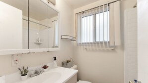 A bathroom with a white sink, mirror, toilet, and shower. There is a window with vertical blinds and a small shelf with toiletries.