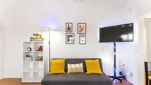 A living room with a grey sofa, yellow cushions, a white shelving unit, and a flat-screen TV mounted on the wall.