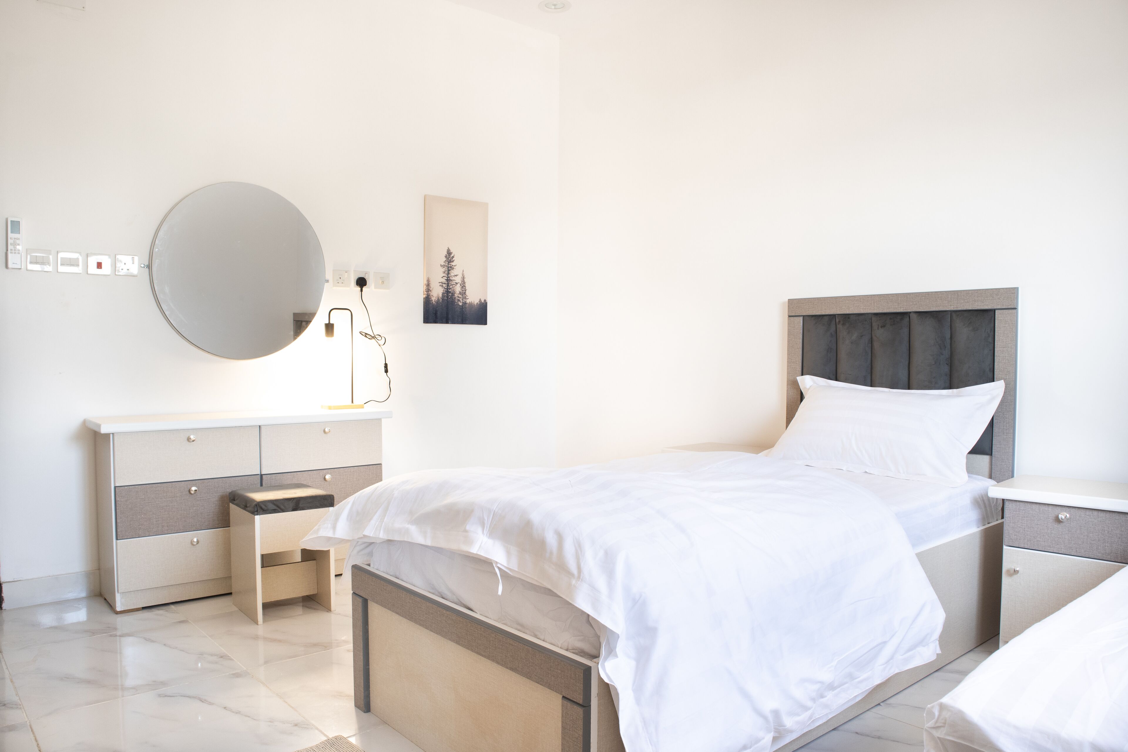 A modern bedroom with a large bed, bedside tables, a dresser, a round mirror, and a wall-mounted lamp.
