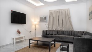 A modern living room with a grey sofa, a black coffee table, a white side table, a flat-screen TV mounted on the wall, and an air conditioning unit.