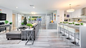 Una sala moderna con sofá, una mesa de centro y televisor. Se observa una zona de cocina con barra y taburetes.