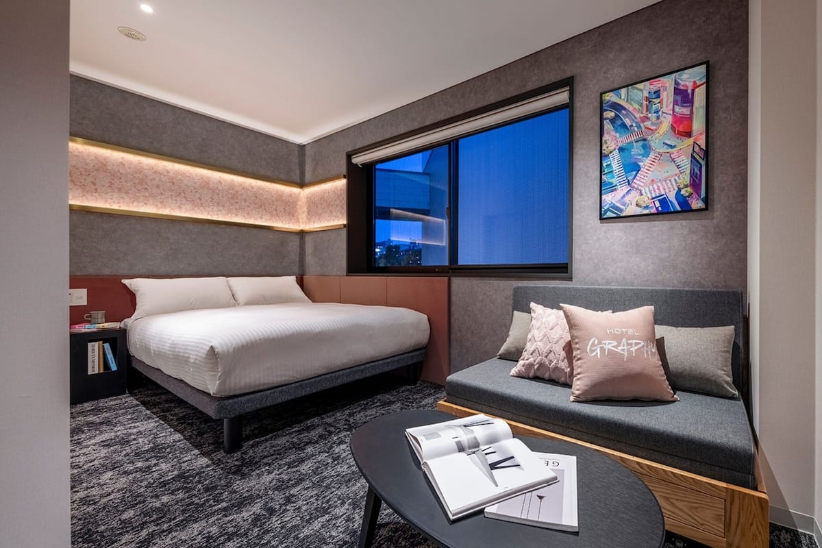 A modern hotel room with a bed, a sofa, a coffee table with magazines, a large window, and a colorful painting on the wall.