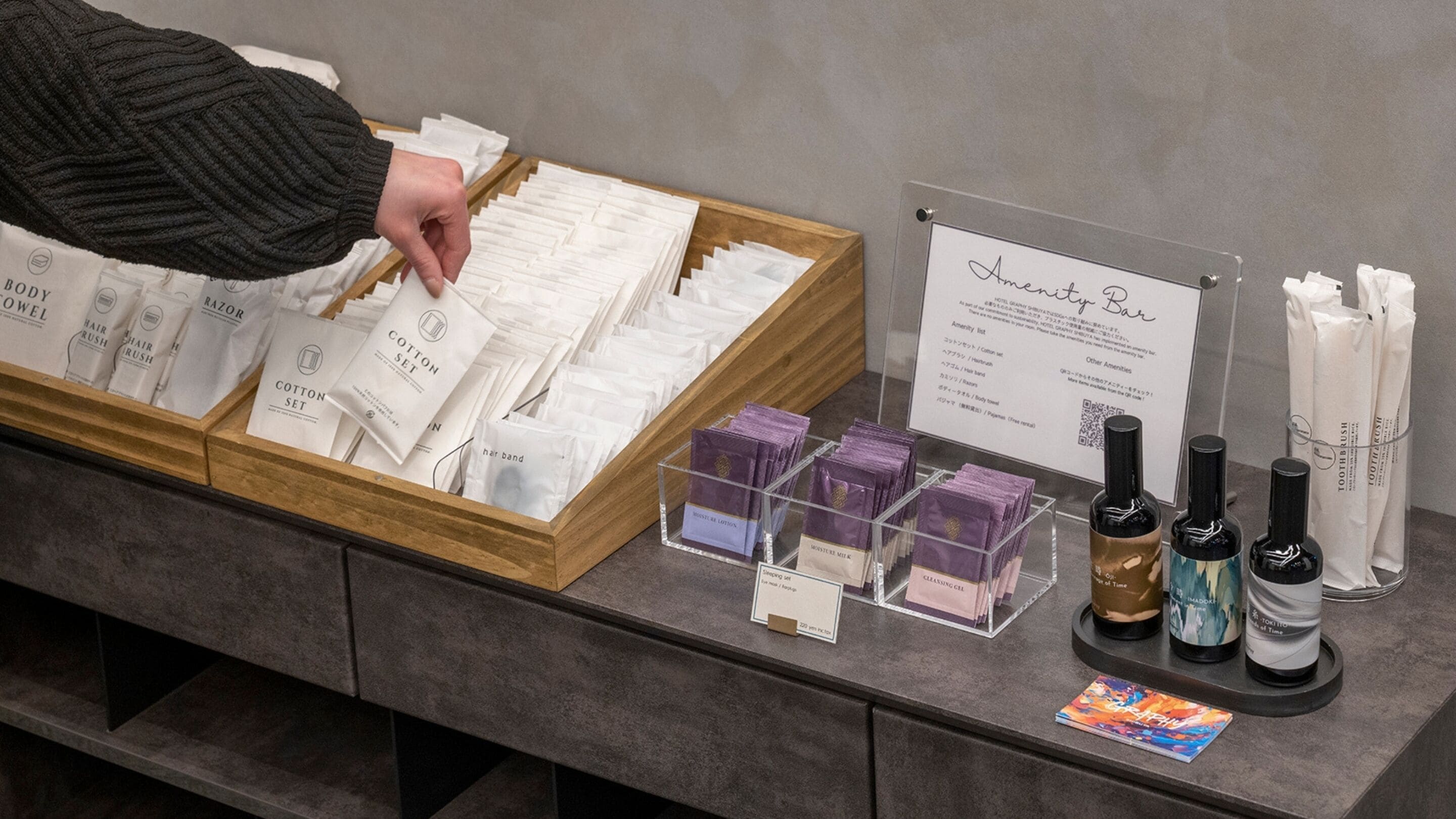 Ein Hotel-Rezeptionsbereich mit einer Holztresen, auf dem verschiedene Pflegeprodukte ausliegen, und einem Schild für die Bar 'Amity Bar'.