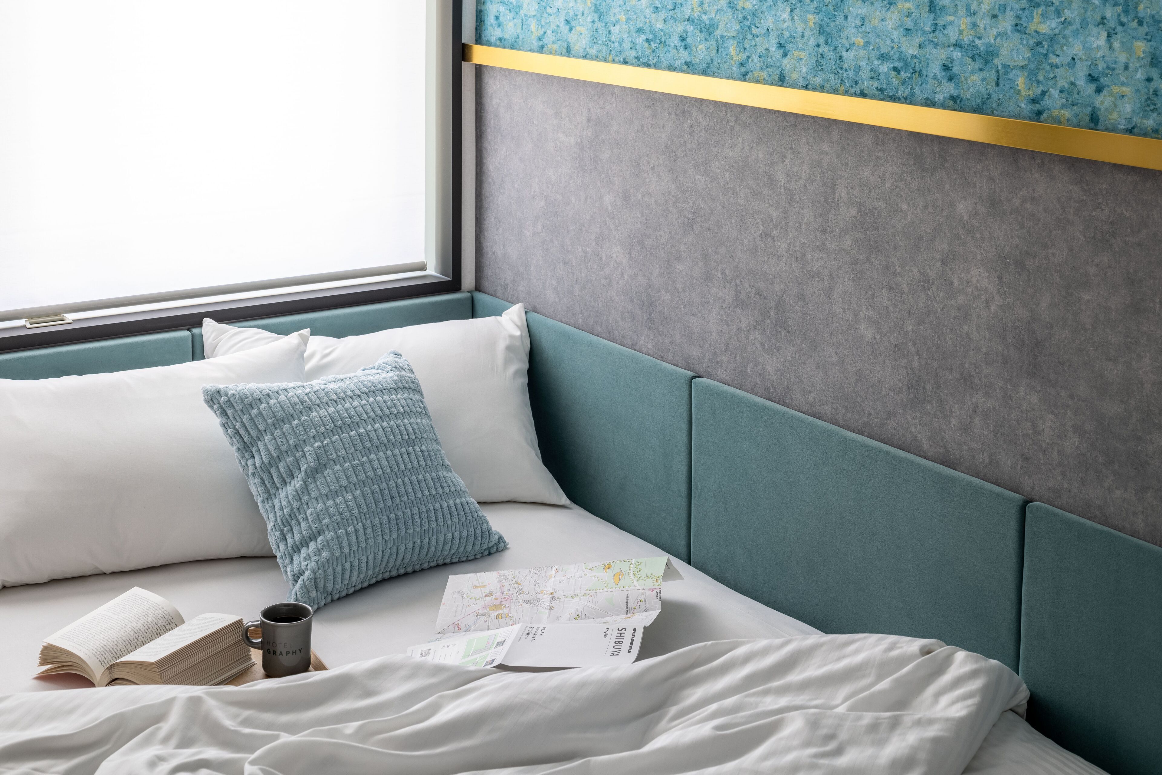 A neatly made bed with a book, a mug, and a map, a window with a view, and a teal headboard.