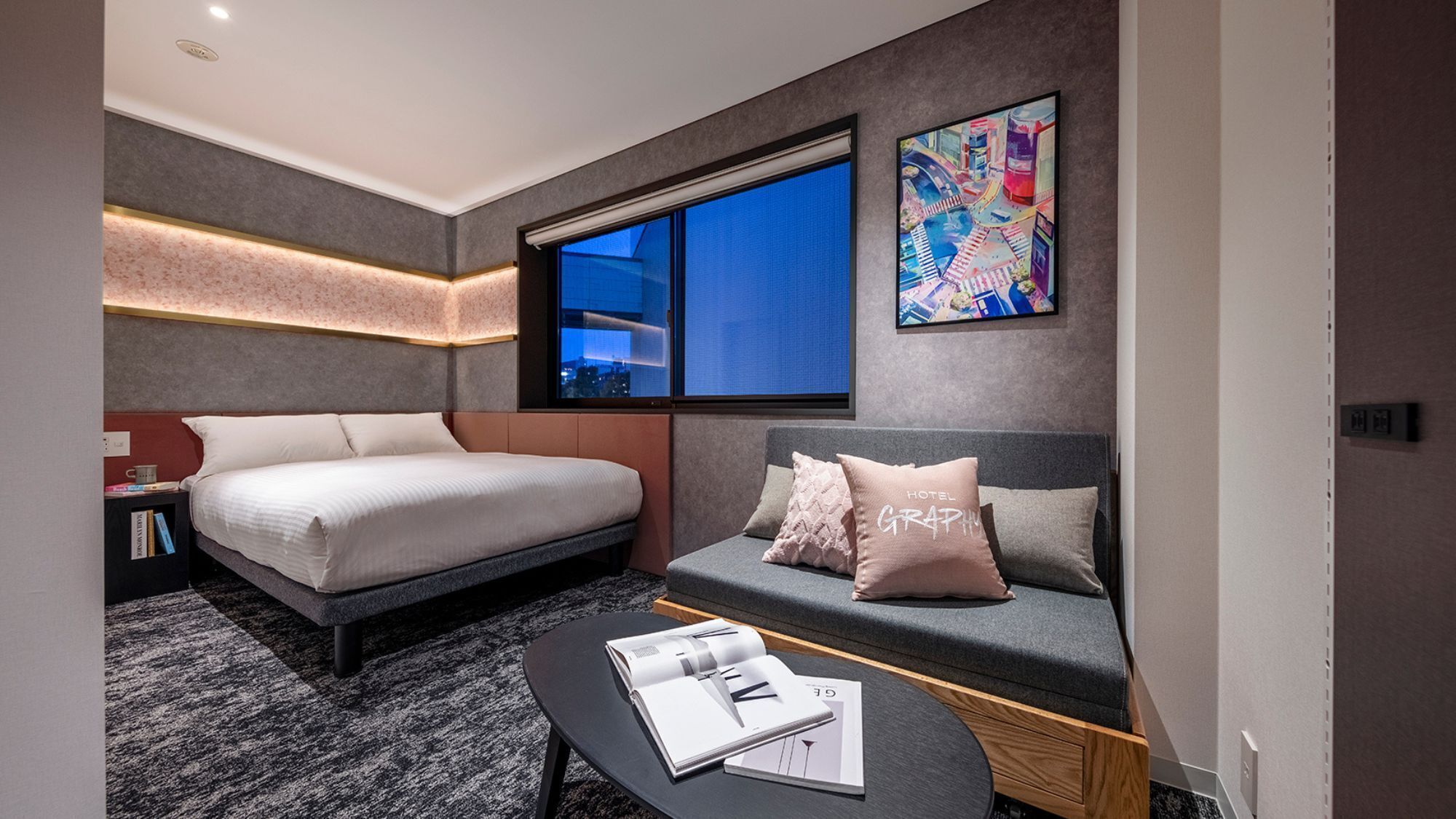 A modern hotel room with a bed, a sofa, a coffee table with magazines, a large window, and a cityscape view.