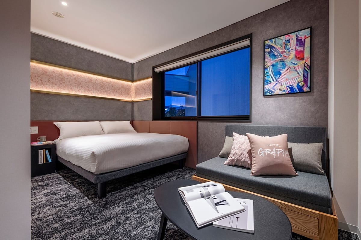Une chambre dâhĂŽtel moderne Ă©quipĂ©e dâun lit, dâun canapĂ©, dâune table basse avec des magazines, dâune grande fenĂȘtre et offrant une vue sur le paysage urbain.