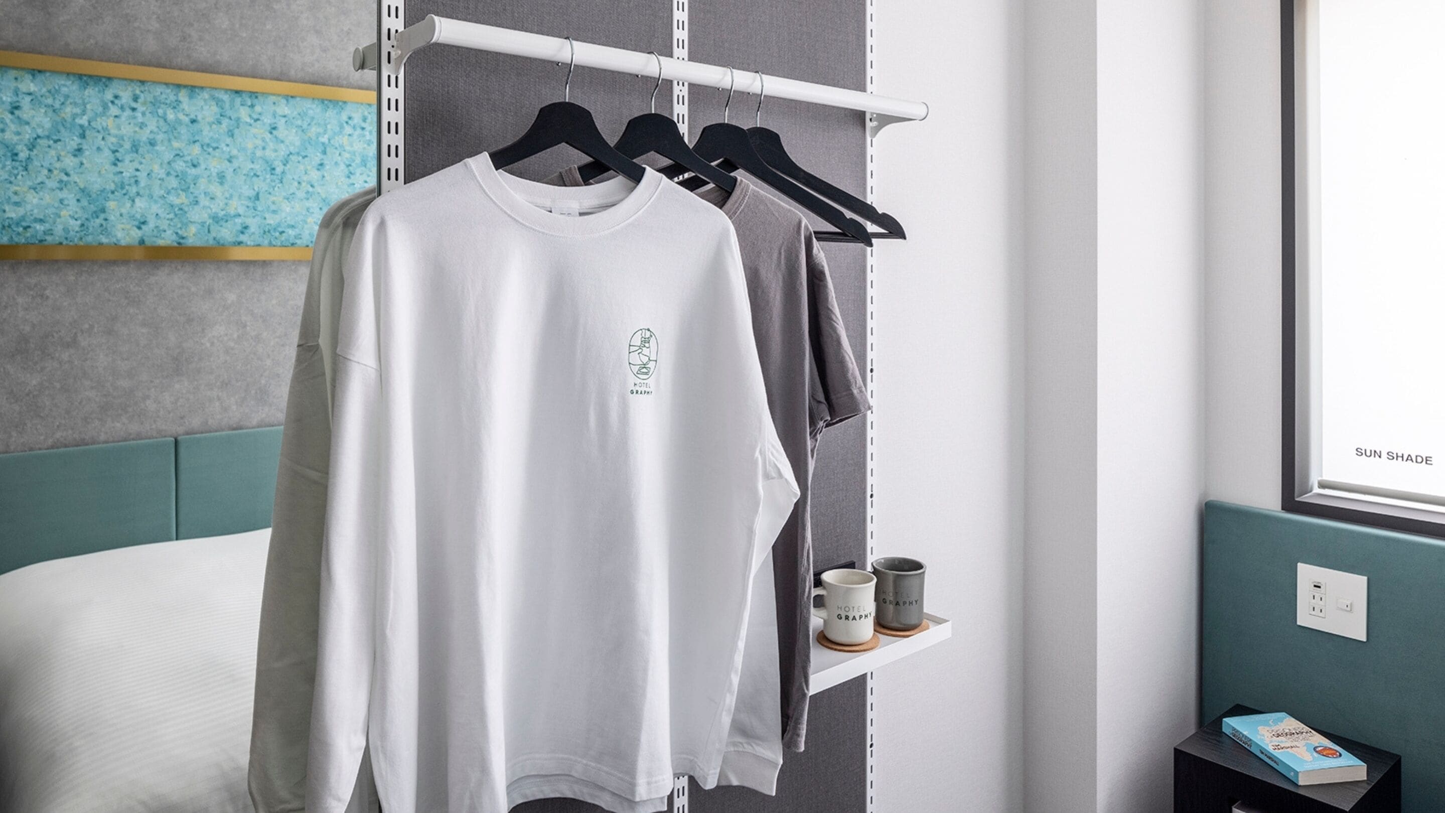 A hotel room with a bed, a shelf with two mugs, and a window with a view of the sky.