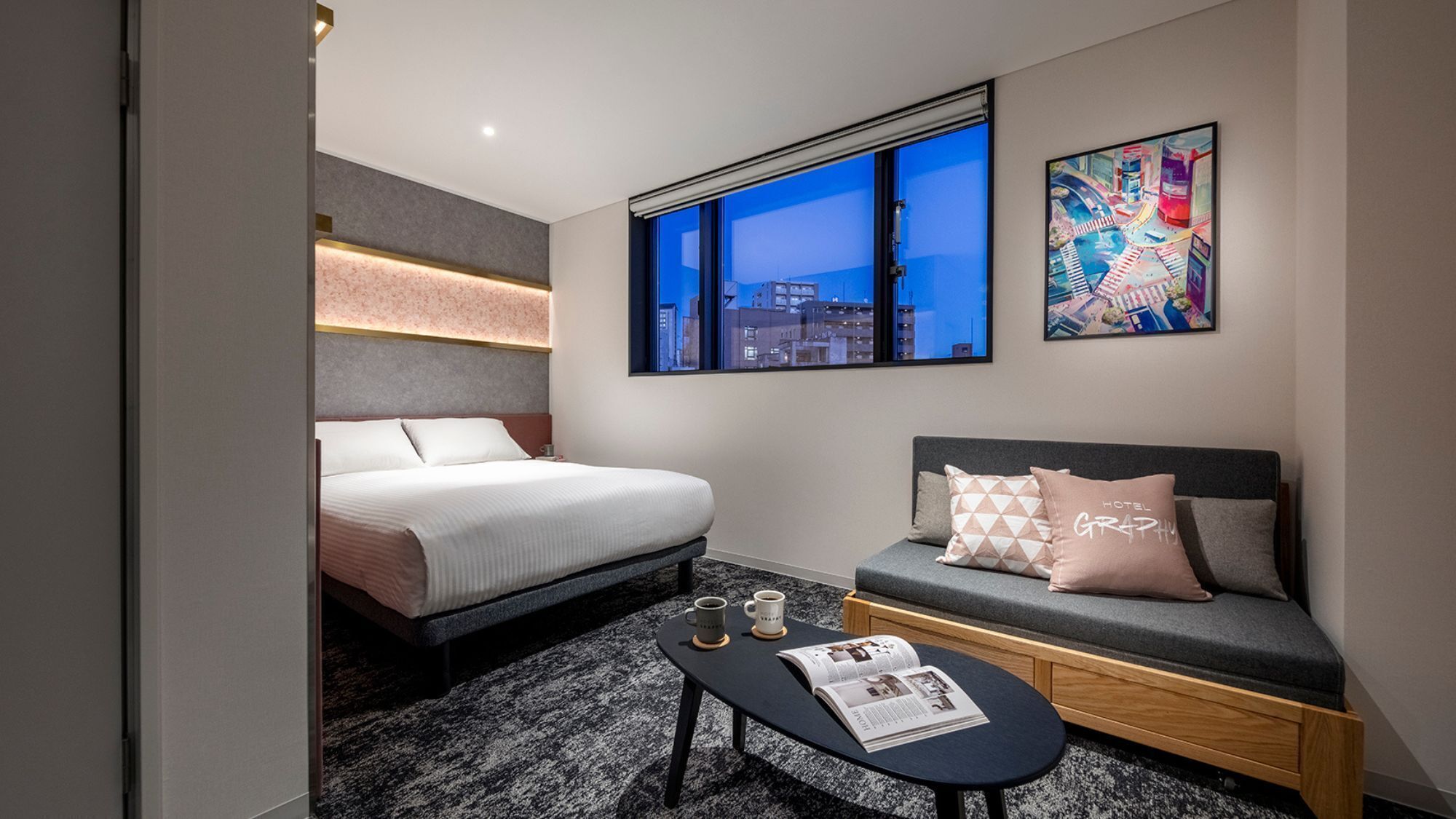A hotel room with a bed, a sofa, a small table with a magazine, and a window with a cityscape view.