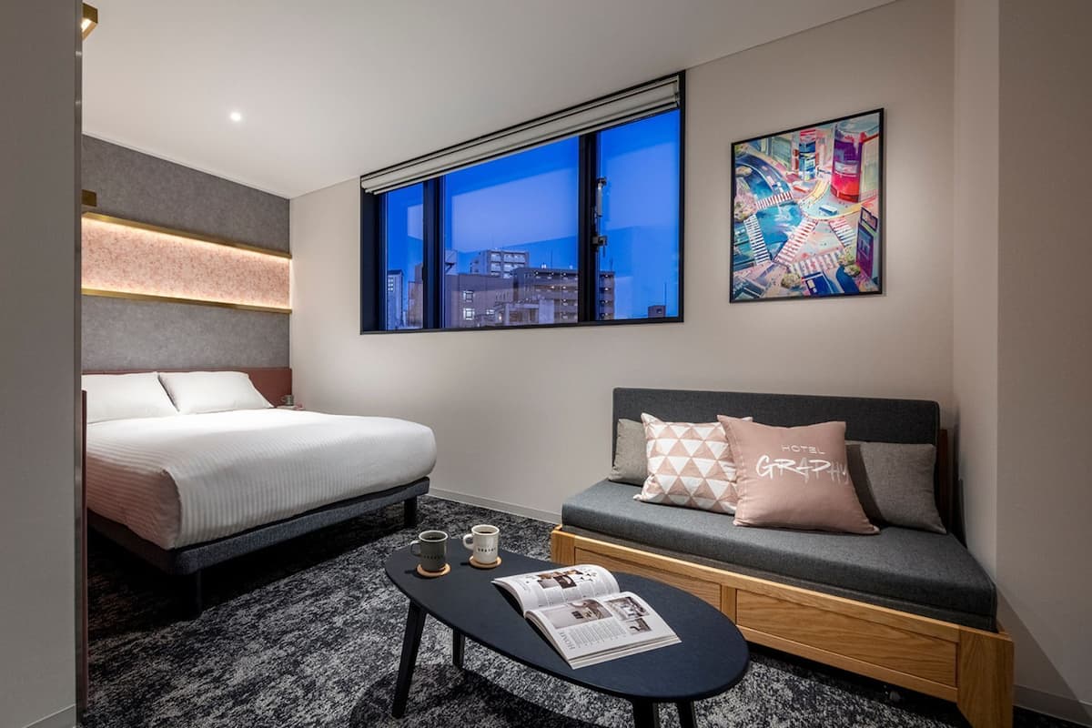 A hotel room with a bed, a sofa, a small table with a magazine, and a window with a cityscape view.