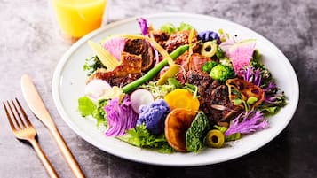 Un plato de comida con diversos vegetales y carnes, acompañado de un tenedor y cuchillo sobre una superficie oscura.