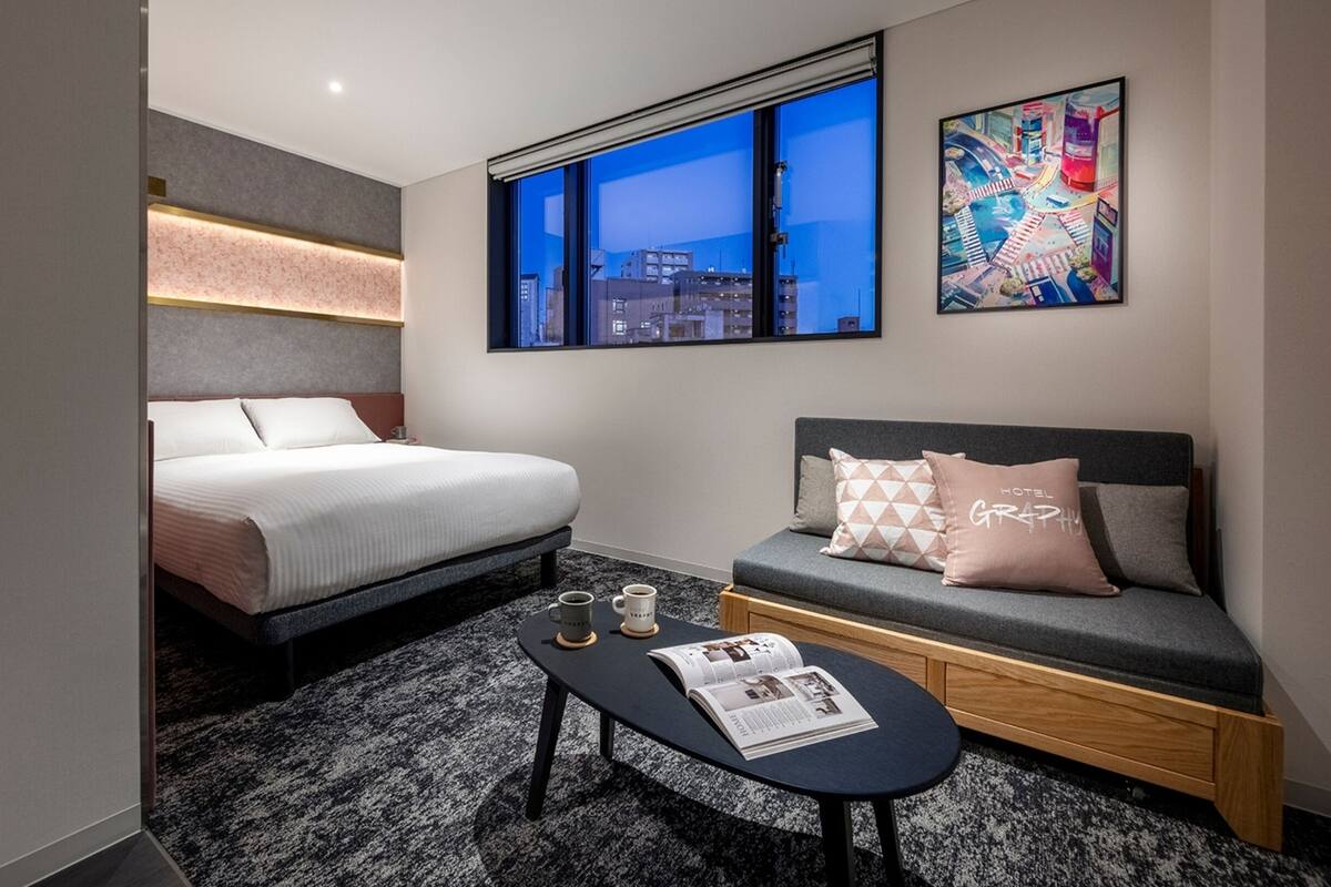 A modern hotel room with a bed, a sofa, a coffee table, and a window with a city view.