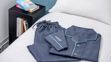 A neatly made bed with a striped white and gray duvet cover. On the bed, there are two dark blue pajama sets with light blue piping and a drawstring. To the left, a black nightstand holds a book titled "NIKKI MONSTER" and a small blue box.
