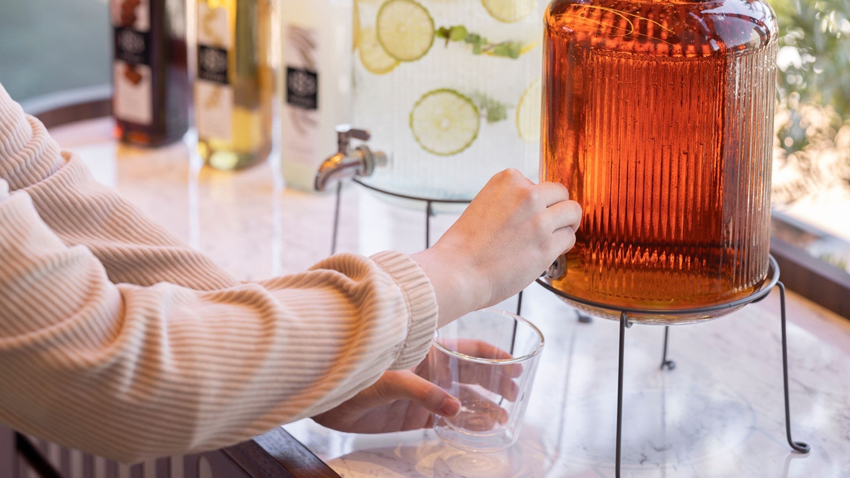 Une personne verse une boisson d’un grand pichet de couleur ambrée dans un verre posé sur un comptoir en marbre.