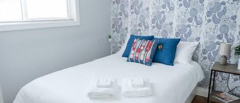 Cama bem arrumada com lençóis brancos e duas almofadas azuis, uma mesinha de cabeceira com abajur, e uma janela com persianas.