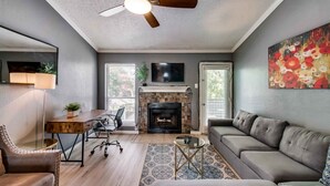 A living room with a fireplace, a ceiling fan, a sofa, a coffee table, and a TV mounted on the wall. - I18 - Apt|Yard|11mi to Fair Park|Sleeps 4 (Irving)