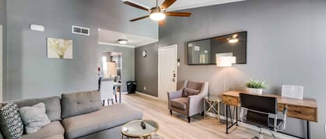 A modern living room with a grey sofa, a round coffee table, a desk with a chair, a ceiling fan, and a large mirror.