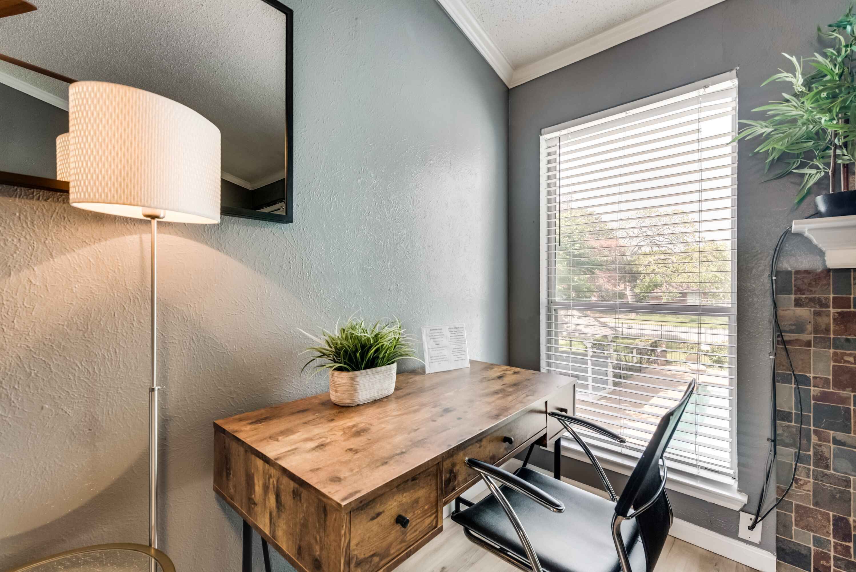 A modern interior with a wooden desk, a black chair, a floor lamp, and a window with blinds.