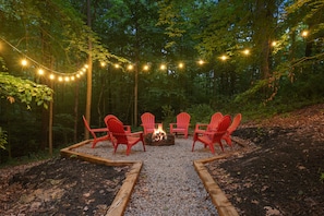 A forest setting with a fire pit and red chairs arranged in a circle, surrounded by a gravel area and wooden borders.