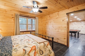 Una habitación con paneles de madera, una cama, un ventilador de techo, una ventana con cortinas y una mesa de billar.