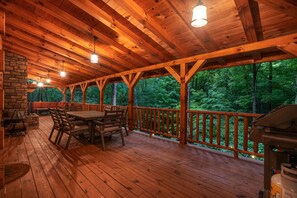 A wooden deck with a stone fireplace, outdoor seating, and a view of a forest.