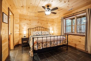 A wooden cabin bedroom with a bed, two bedside tables, a ceiling fan, and a window with curtains.